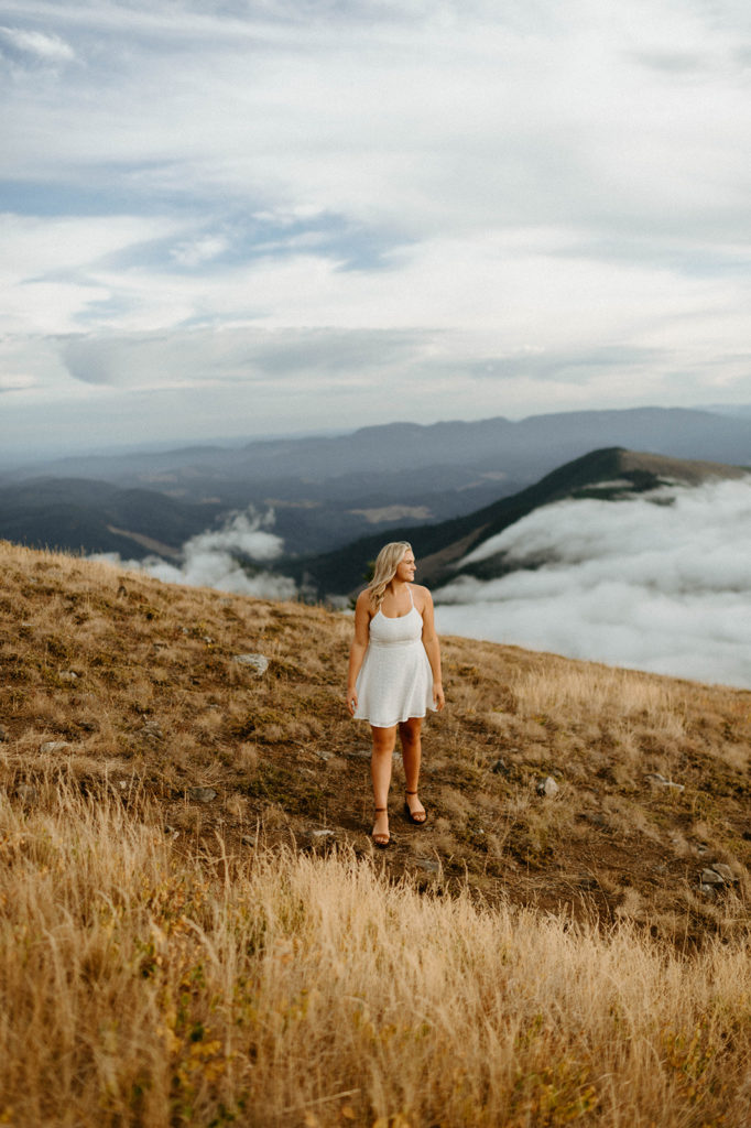 senior in white dress standing on grassy hillside 