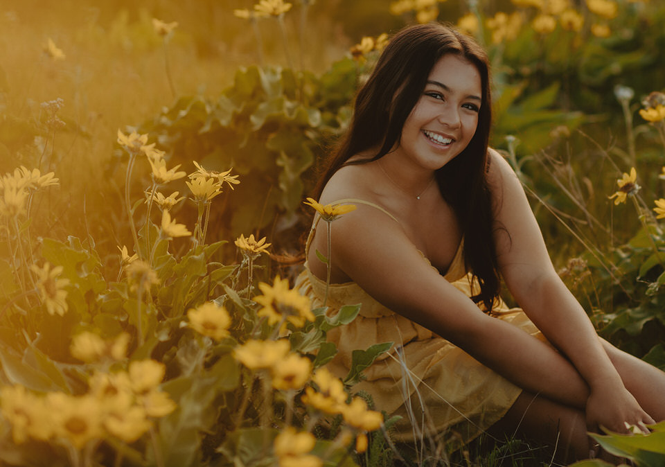 Close-up of a smiling woman sitting among sunflowers at sunset