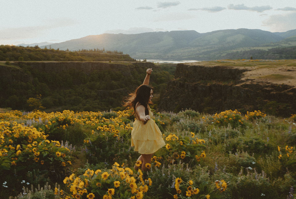 Woman joyfully dancing in a field of sunflowers with a mountainous backdrop at sunset