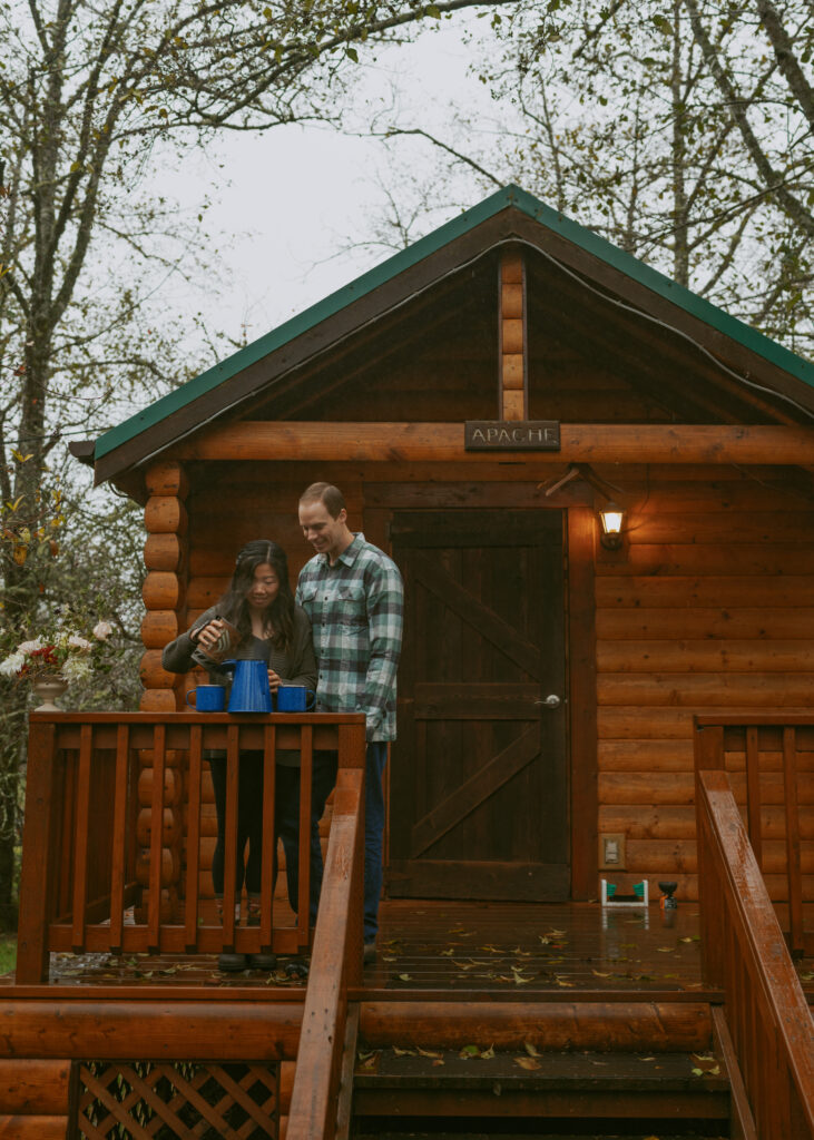 Girl pouring hot water from a tumbler to the blue pot sitting on the railings of a wood cabin with her partner