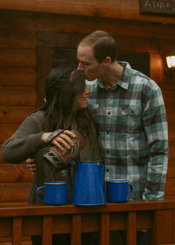 Girl pouring hot water from a tumbler to the blue pot sitting on the railings of a wood cabin with her partner kissing her forehead