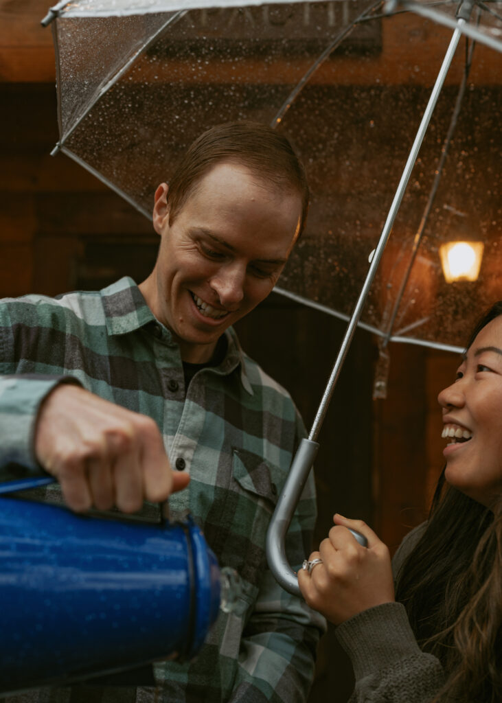 Boy pouring the hot drink from the blue pot to the blue cup while his partner is happily looking at him and holding a clear umbrella for the both of them