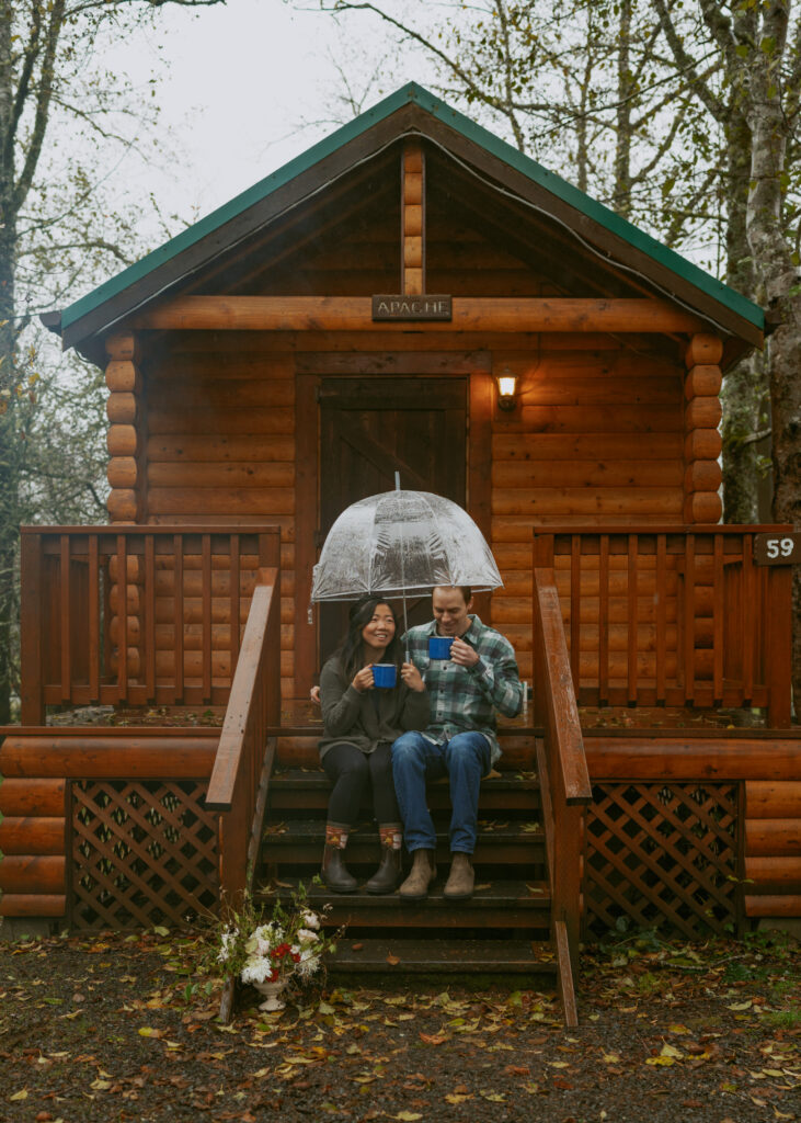 Couple sitting on a wooden stairs of a cabin while drinking coffee from blue cups and holding a clear umbrella