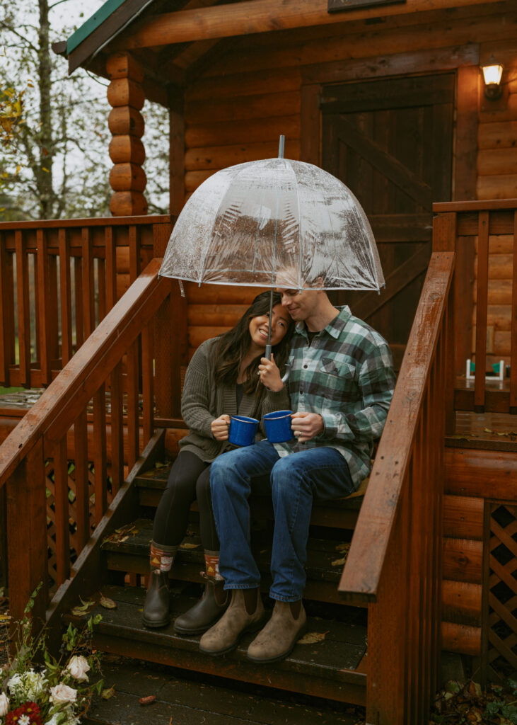 Couple sitting on a wooden stairs of a cabin while drinking coffee from blue cups and holding a clear umbrella