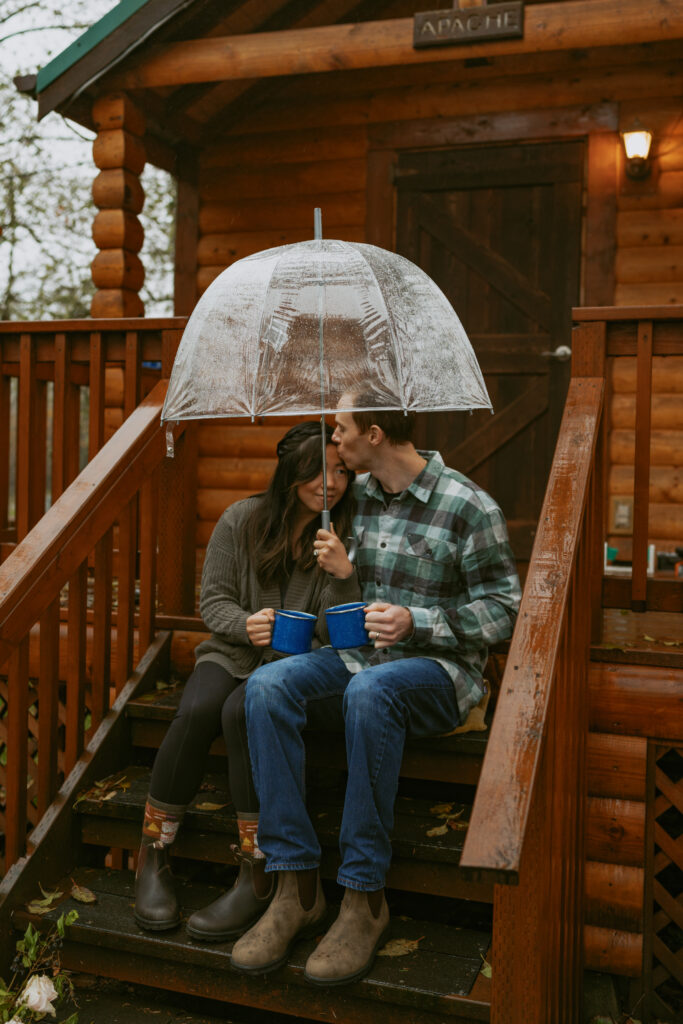 Couple sitting on a wooden stairs of a cabin while each of them holds their blue cups while the boy is kissing the forehead of his partner who's holding a clear umbrella