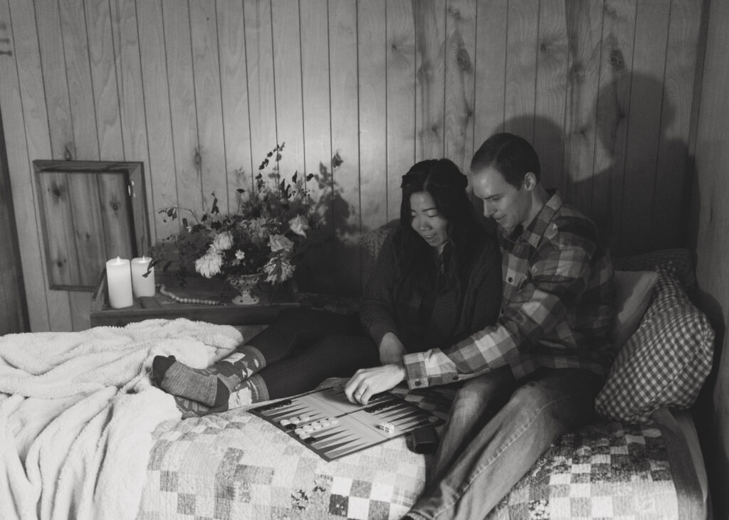 couple enjoys a cozy moment playing backgammon on a quilt-covered bed in a rustic wooden room, surrounded by candles and flowers