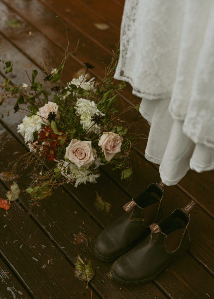 Dreamy Elopement scene featuring a bouquet of blush roses and white flowers beside dark leather shoes on a wooden floor, with a glimpse of a lace bridal gown