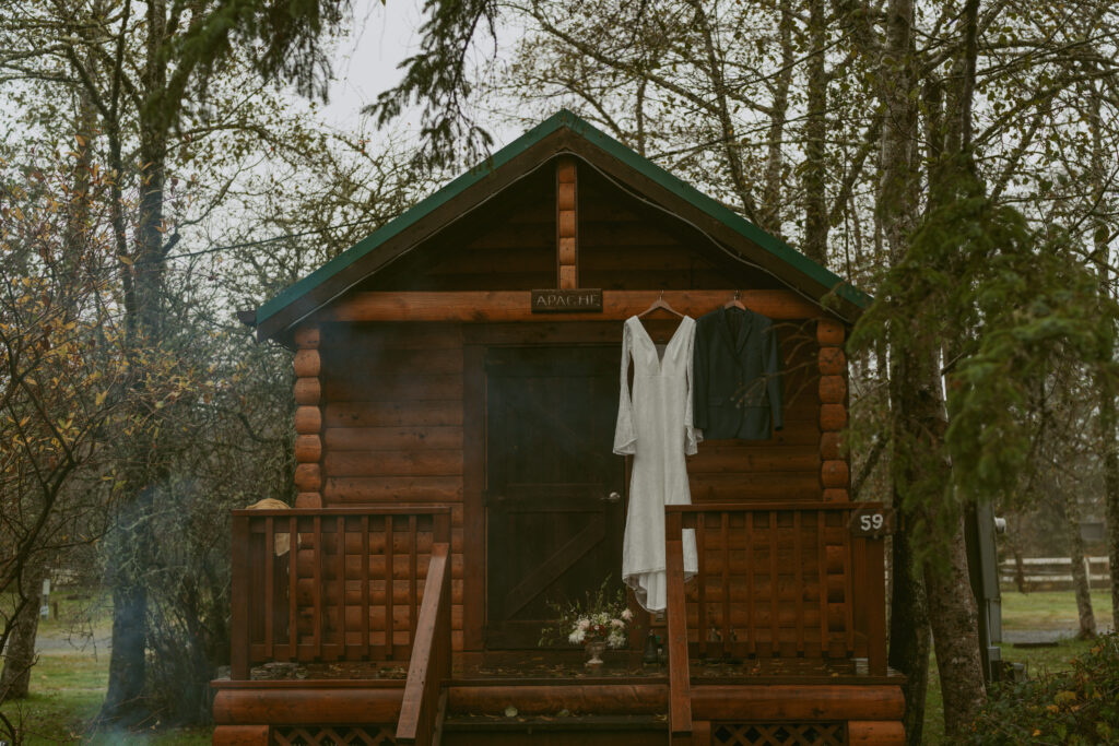 A dreamy elopement scene with a bridal gown and a suit hanging outside a wooden cabin and a small bouquet on the porch
