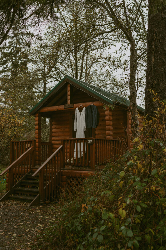 A dreamy elopement scene at Cannon Beach with a bridal gown and suit hanging on the porch of a rustic wooden cabin surrounded by lush greenery