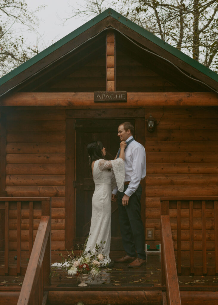 Bride fixing her groom's tie on the porch of a wooden cabin and featuring a floral bouquet for their dreamy elopement
