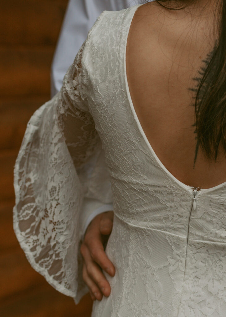 Close-up of a bride in her lace wedding gown featuring the sleeves, with the groom's hand gently resting on her back