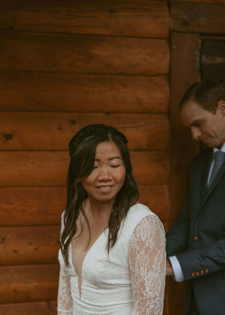 Smiling bride with her eyes closed, dressed in an elegant lace wedding gown and the groom, in a dark suit, fixing the back part of the gown of the bride