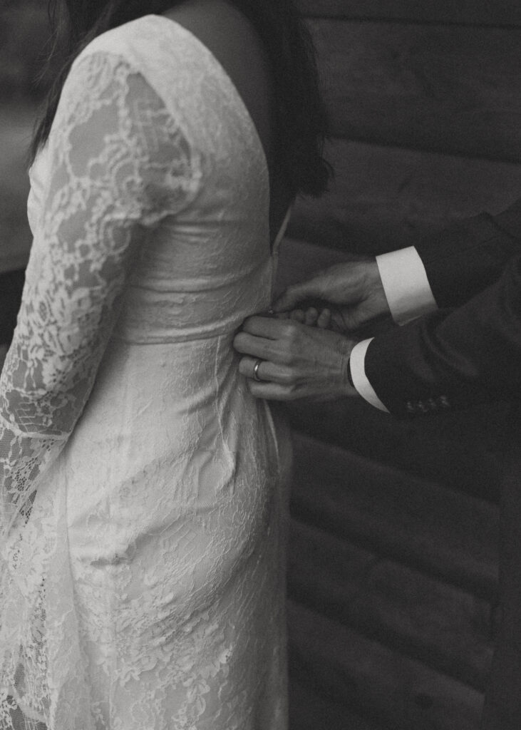 A black and white photo of the groom gently touches the lace-detailed back of his bride's dress
