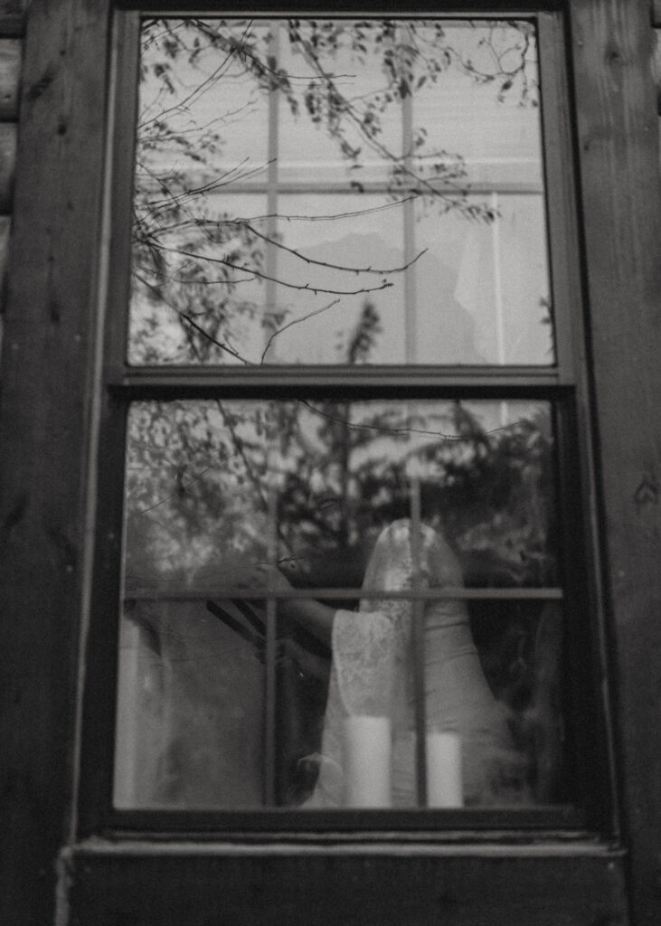 A bride in her wedding gown seen through a rustic window, with the reflections of trees and candles surrounding her