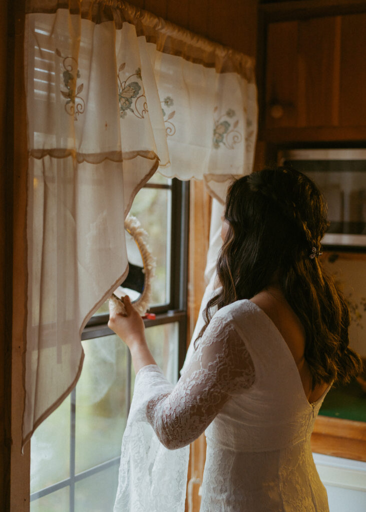 Bride in a lace wedding gown holding hand mirror by a window with delicate floral curtains, in soft natural light