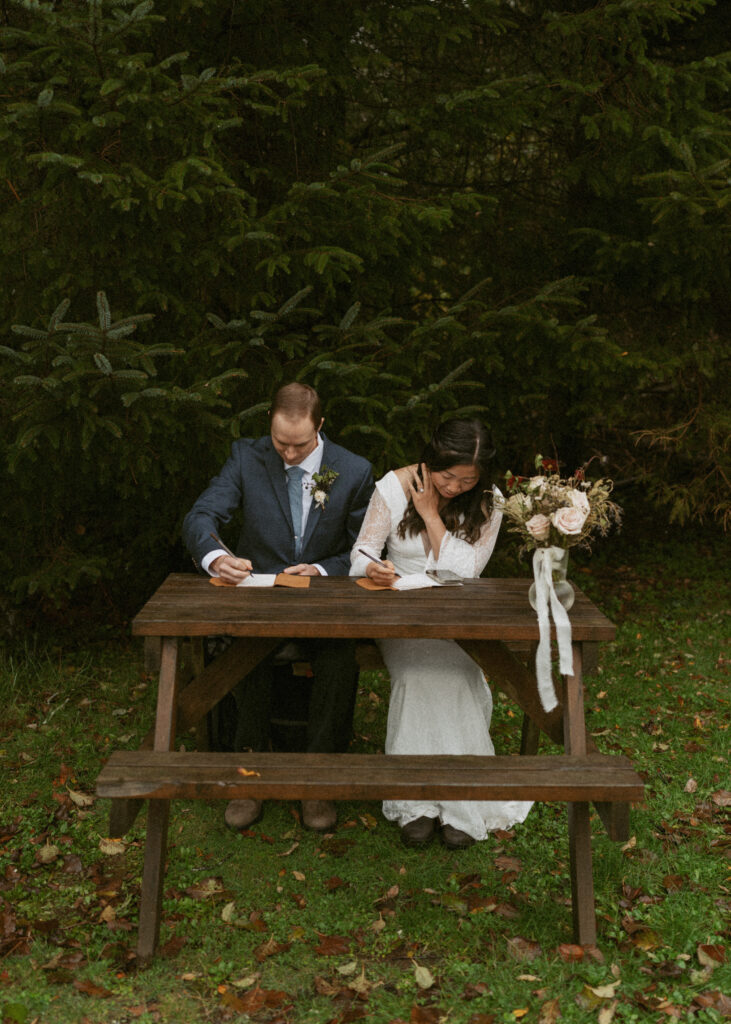 Bride and groom sitting in a wooden table in a forest setting, writing their vows together, with a bouquet of flowers draped over the table's edge