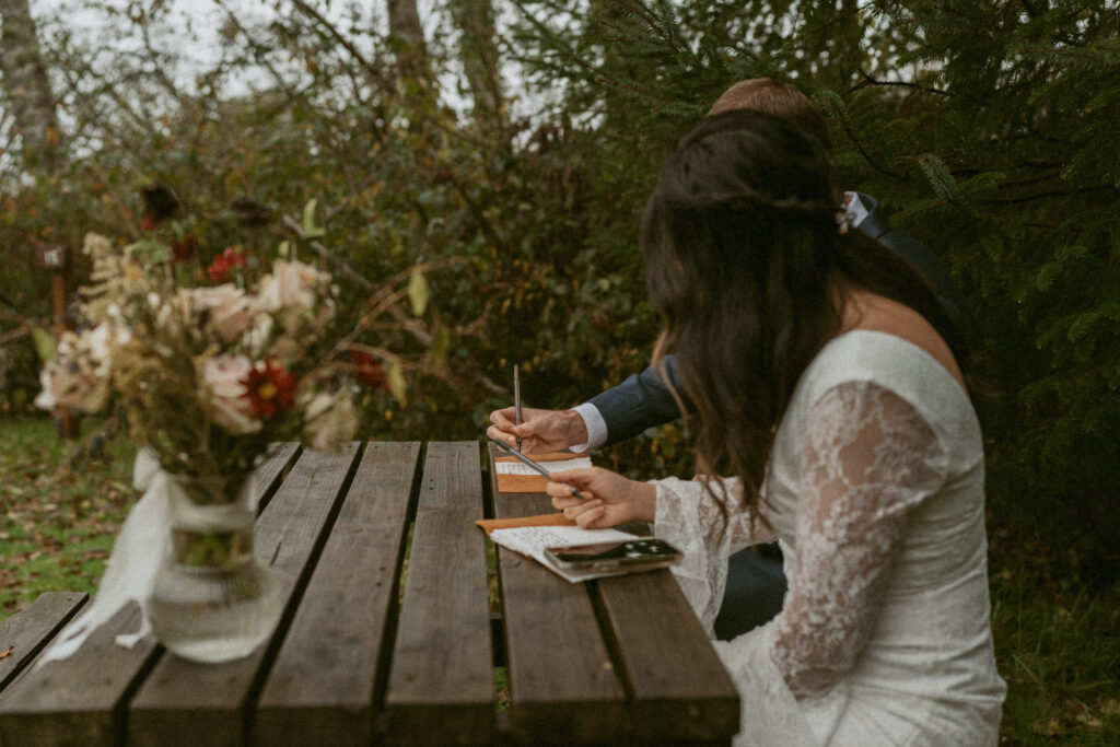 Bride and groom sit side by side at a wooden table outdoors, writing their vows from smartphones to notebooks, surrounded by lush greenery