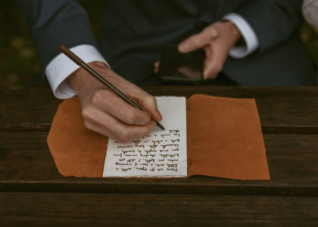 Groom in his suit transfers her vows from a smartphone to a notebook on a wooden table