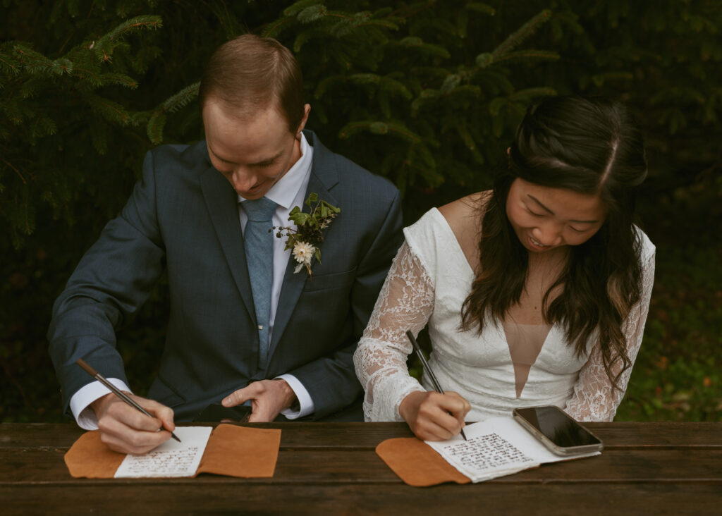 Bride and groom sitting in a wooden table in a forest setting, writing their vows together