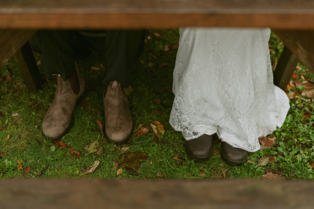 Under a table, a groom's muddy boots sit beside a bride's lace gown and black shoes on a leafy, grassy ground
