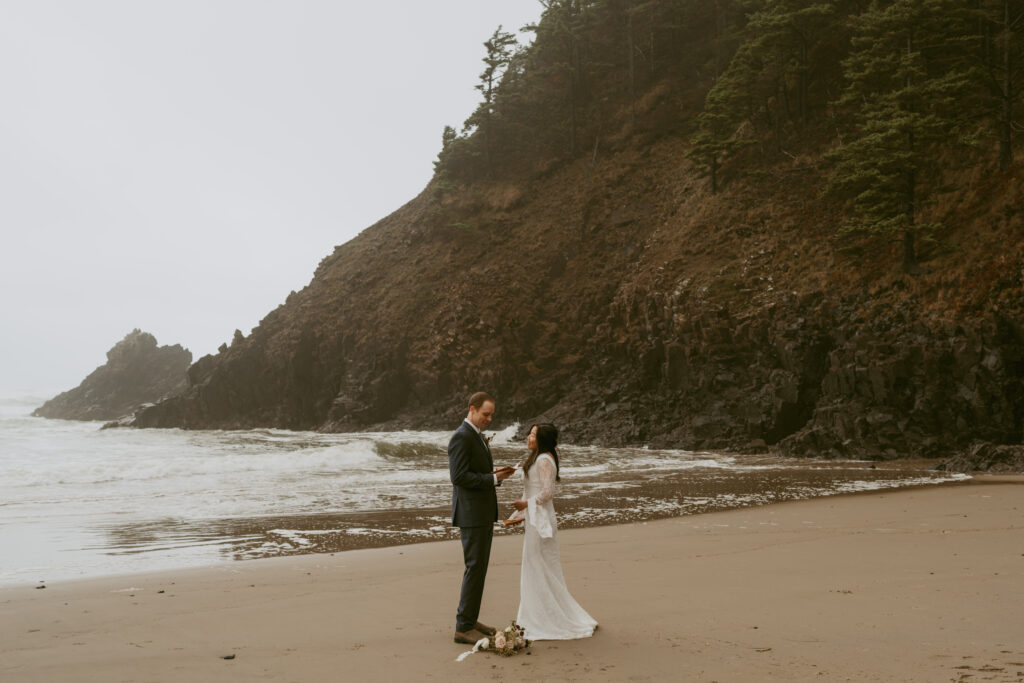 Bride and groom exchange vows on a foggy beach, with a lush, towering cliff in the background