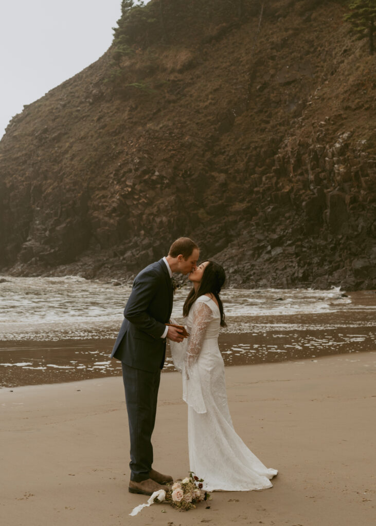 A groom in his suit kisses his bride on a sandy beach, an a cliff in the background