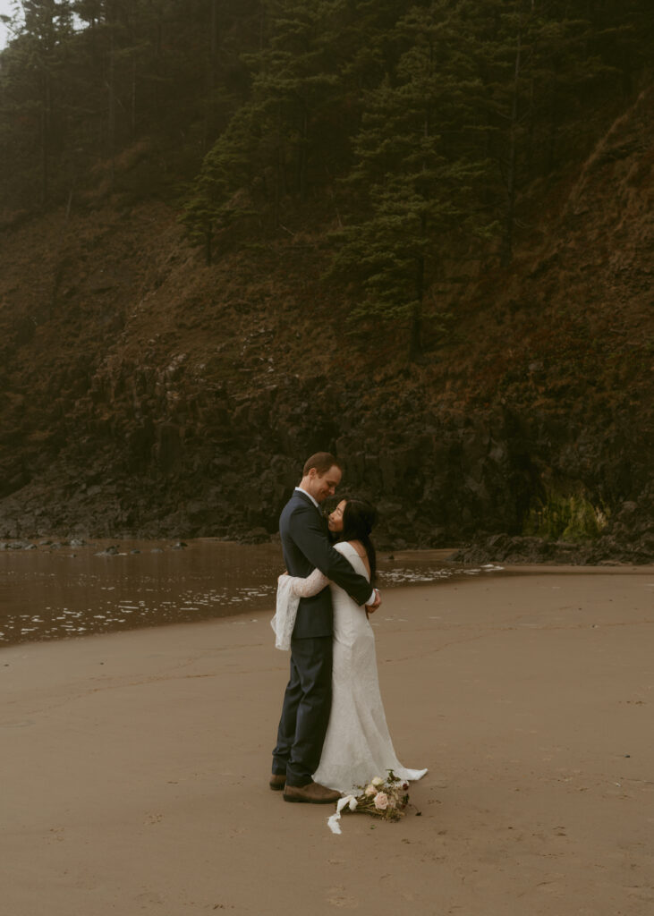 Groom hugging his bride, both smiling beside the beach with crashing waves and cliffs
