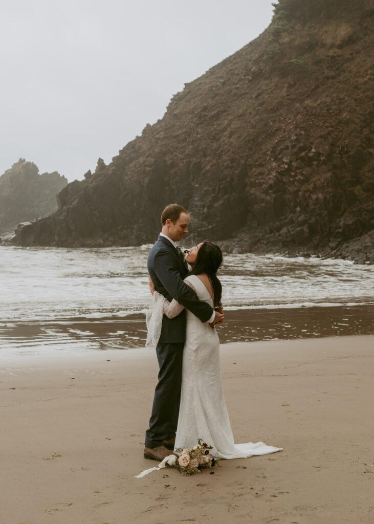 Couple stands closely, looking at each other, surrounded by the scenery of a foggy beach and steep cliffs