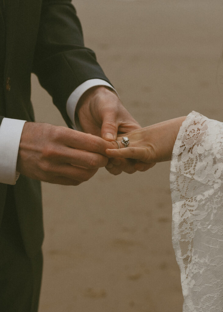 Close-up of a groom placing the wedding ring on the bride's finger, against a sandy background