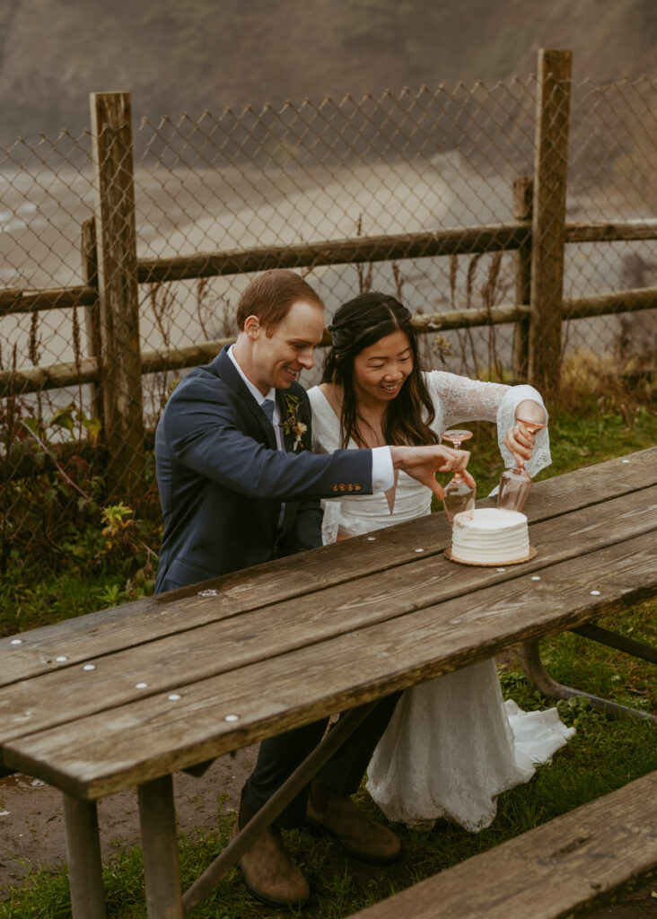 Bride and groom sitting in a wooden table cutting and holding two wine glasses to cut the cake placed on the table