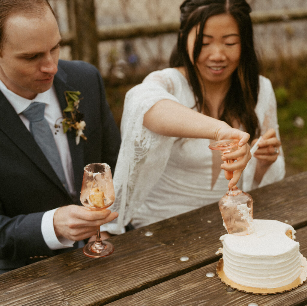 Bride cutting the cake using her glass, while groom holding his glass that has cut cake inside