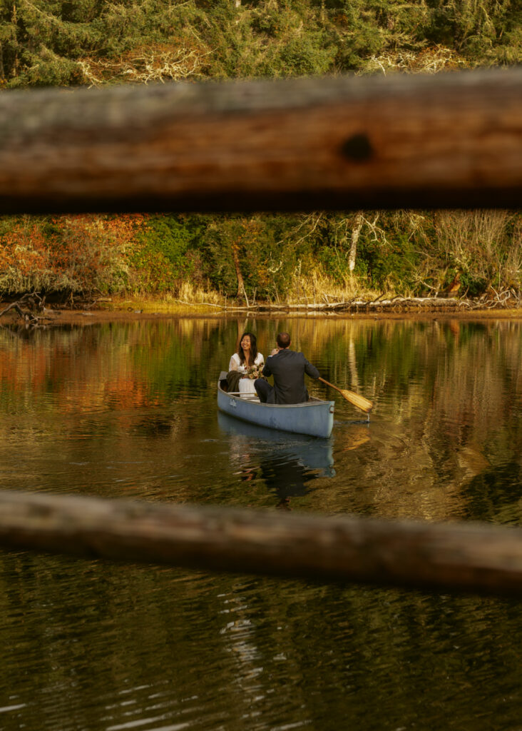 Bride and groom rowing a canoe on a calm lake, framed by wooden fence rails, with autumn-colored trees reflected in the water
