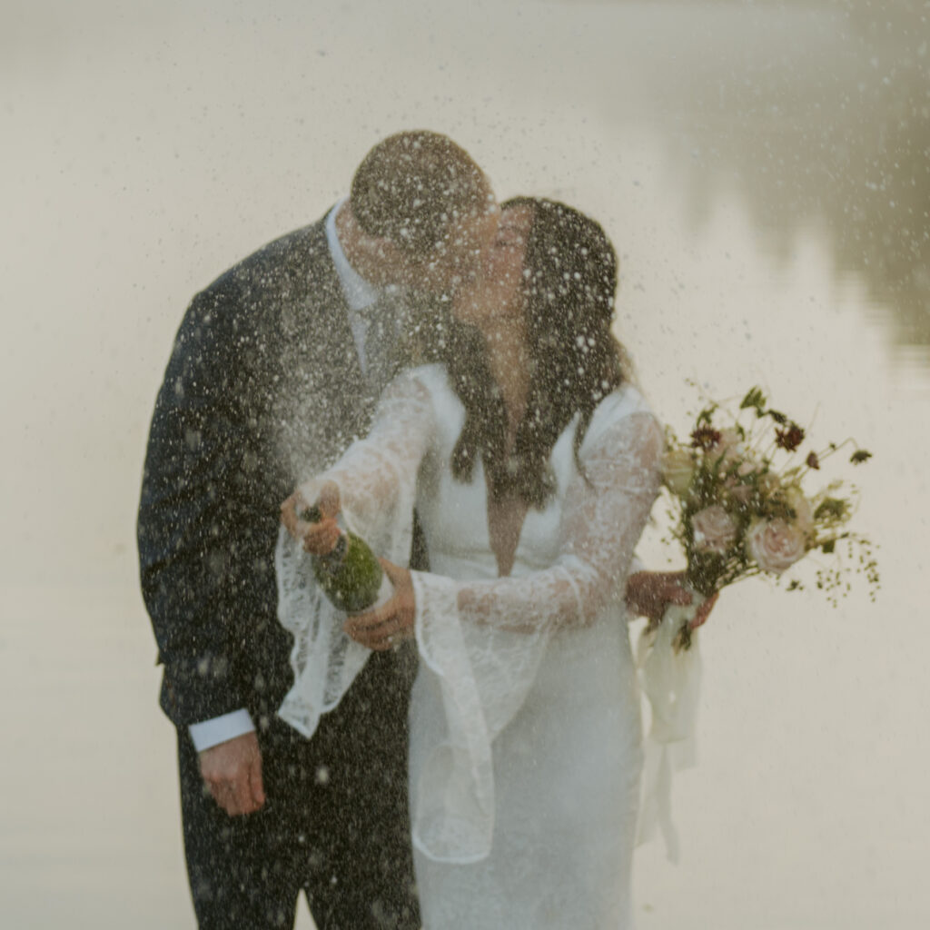 Bride opens a sparkling champagne wine with her groom kissing her on the lakeside