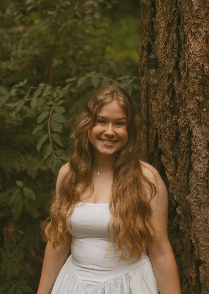 Young woman in her beautiful white dress leaning against the tree and green bushed surrounding it