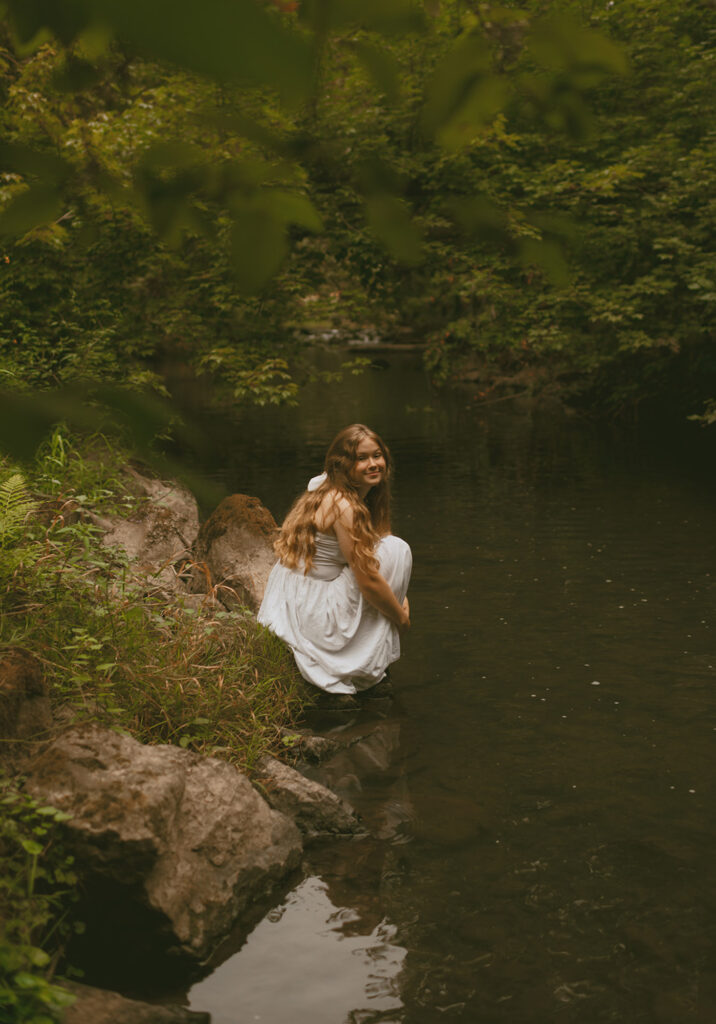 young woman in white dress seated on river stone for her cottage core aesthetic photo session in Oregon