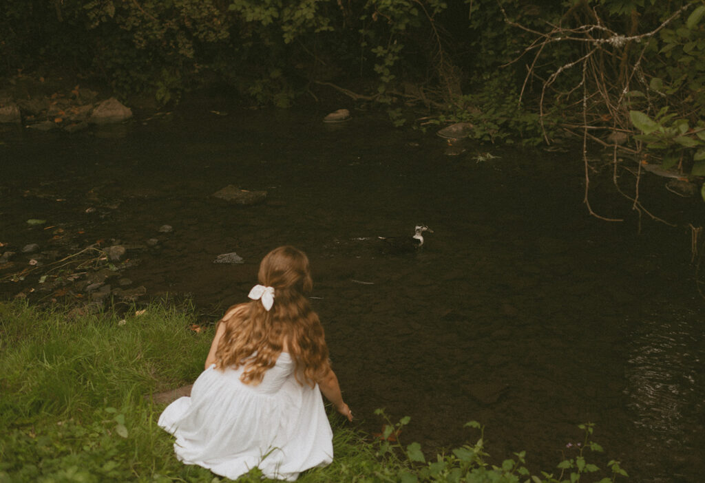 Young woman in white dress seated near river aiming for the duck