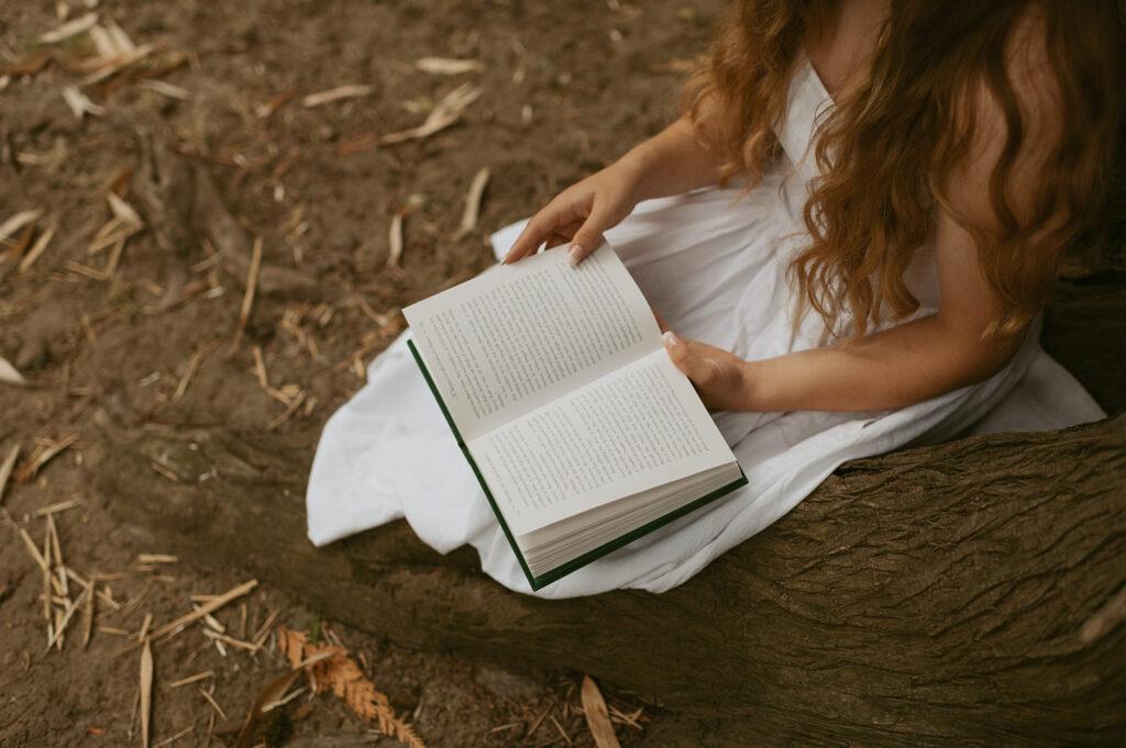 Young woman in a white dress sitting down beside a tree reading a book