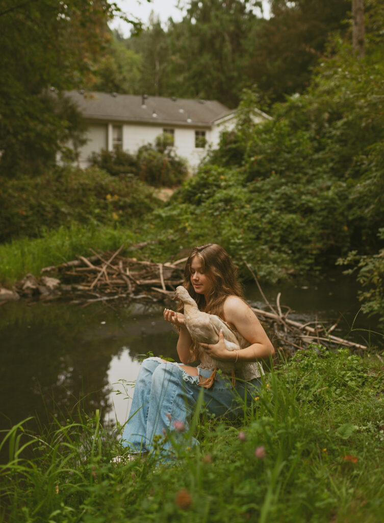 young woman holding a duck by a pond with a house in the background for her cottage core aesthetic photo session in Oregon