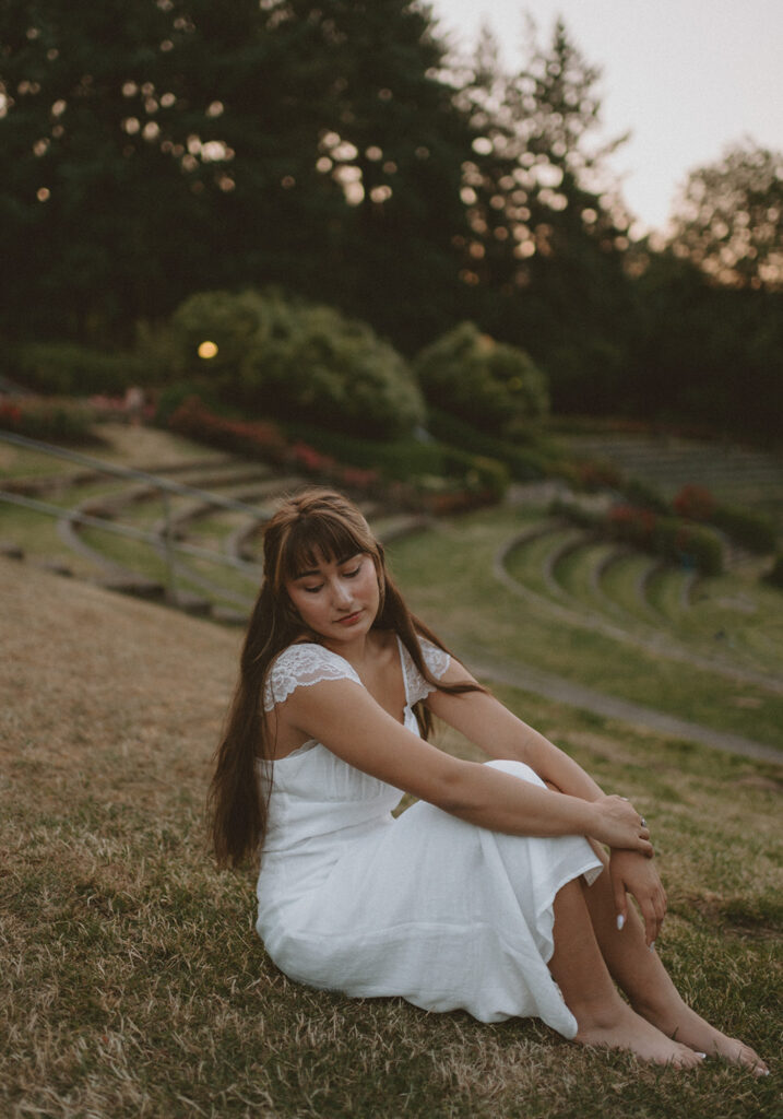 young woman in a beautiful white dress sitting down against the green lawn landscape for her Senior picture session in Portland, Oregon