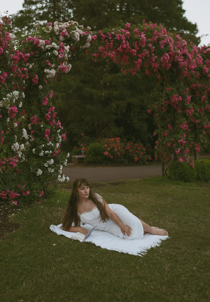 Young woman lying down on a white blanket, dressed in a white and surrounded by towering rose bushes