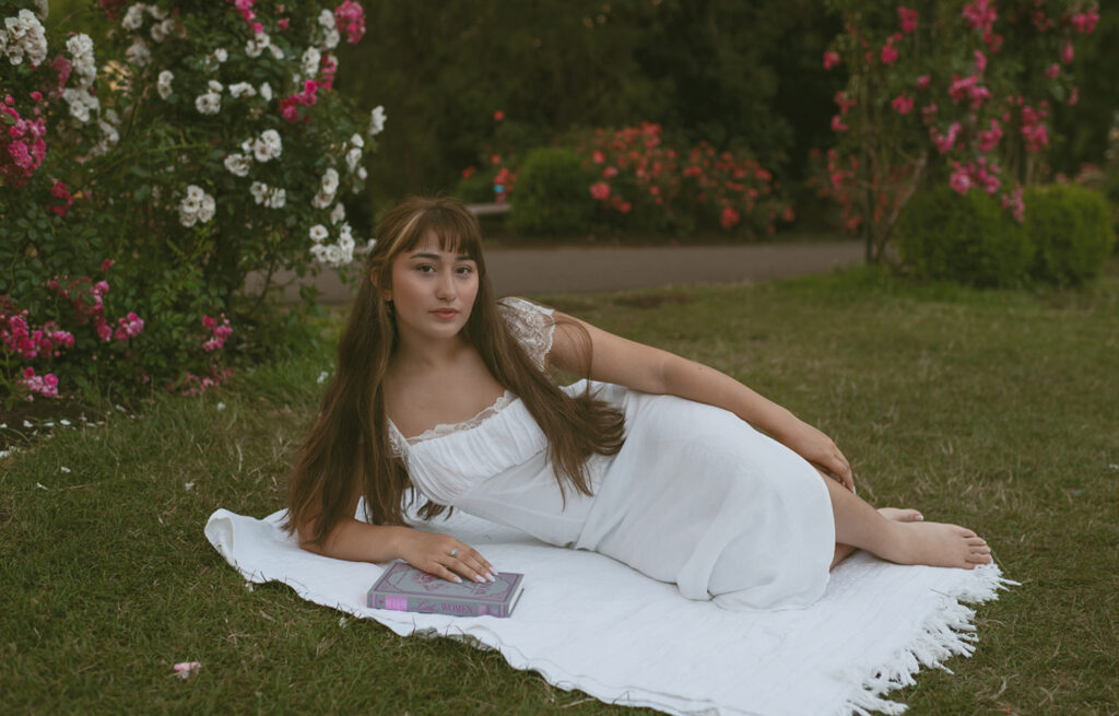 Young woman lying down on a white blanket, holding a book, dressed in a white and surrounded by rose bushes for her Senior picture session in Portland, Oregon