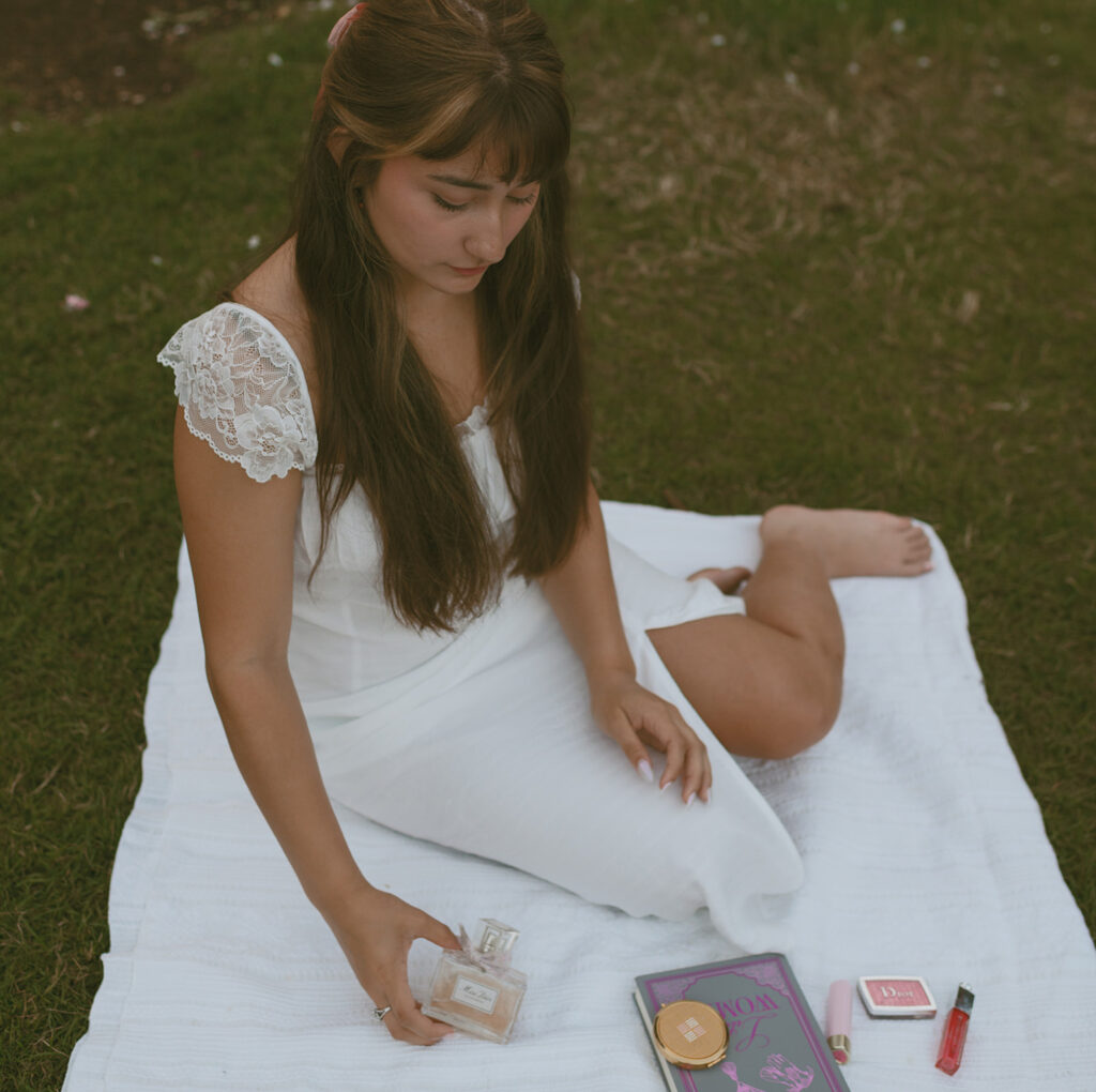 Young woman sitting down on a white blanket, dressed in a white and surrounded by rose bushes for her Senior picture session in Portland, Oregon