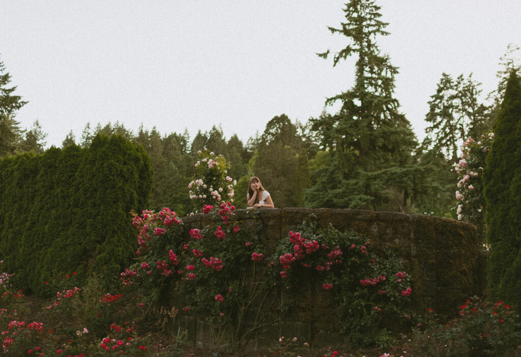 Young woman leaning on a stone wall surrounded by lush rose bushes and tall trees in the background