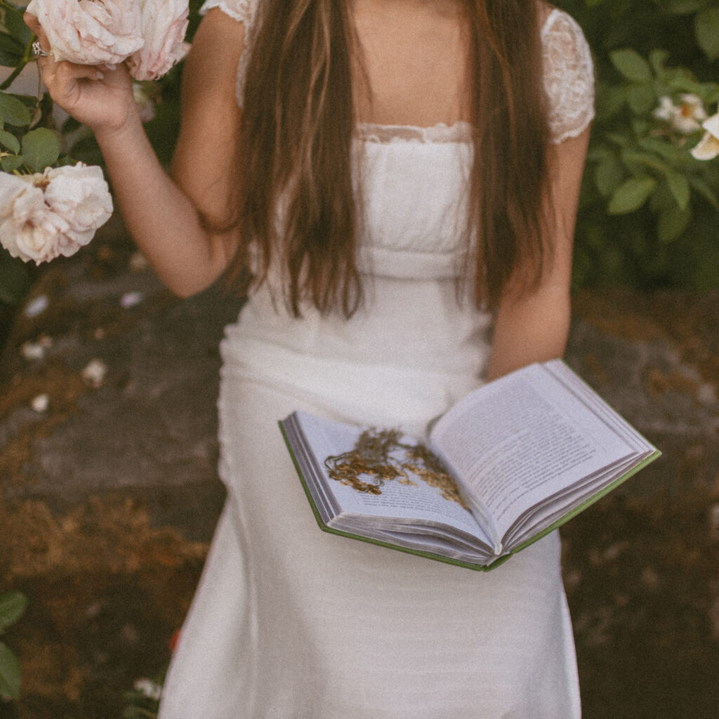 Young woman wearing a white dress sitting down on a rock by the bushes holding her book that has dried flowers