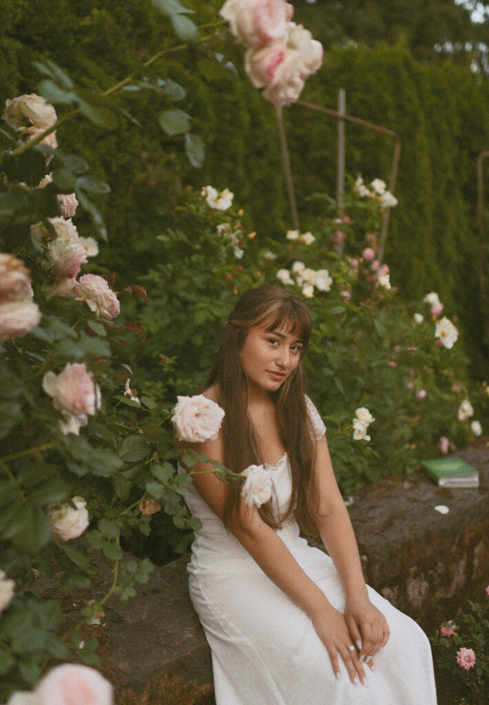 young woman sitting on a rock by rose bushes in a white dress