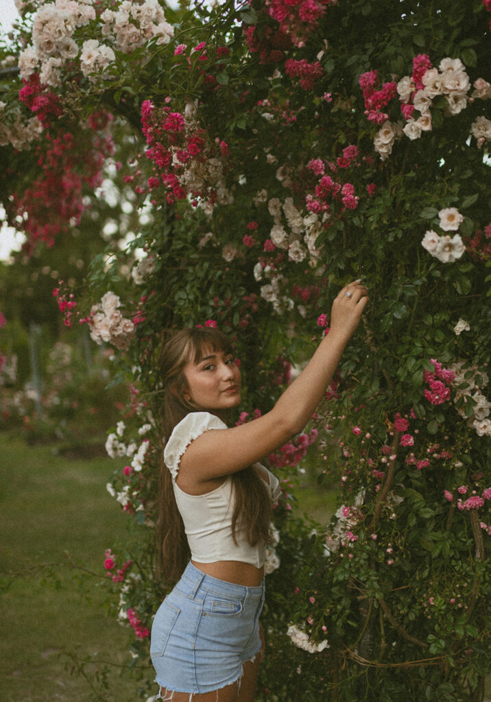Young woman standing while reaching out to touch some high pink roses, dressed in a white crop top and denim shorts