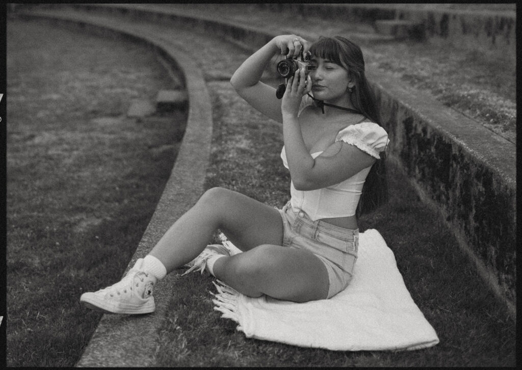Black and white photo of a young woman sitting down on a white blanket on a landscaped grass steps taking a picture using her camera