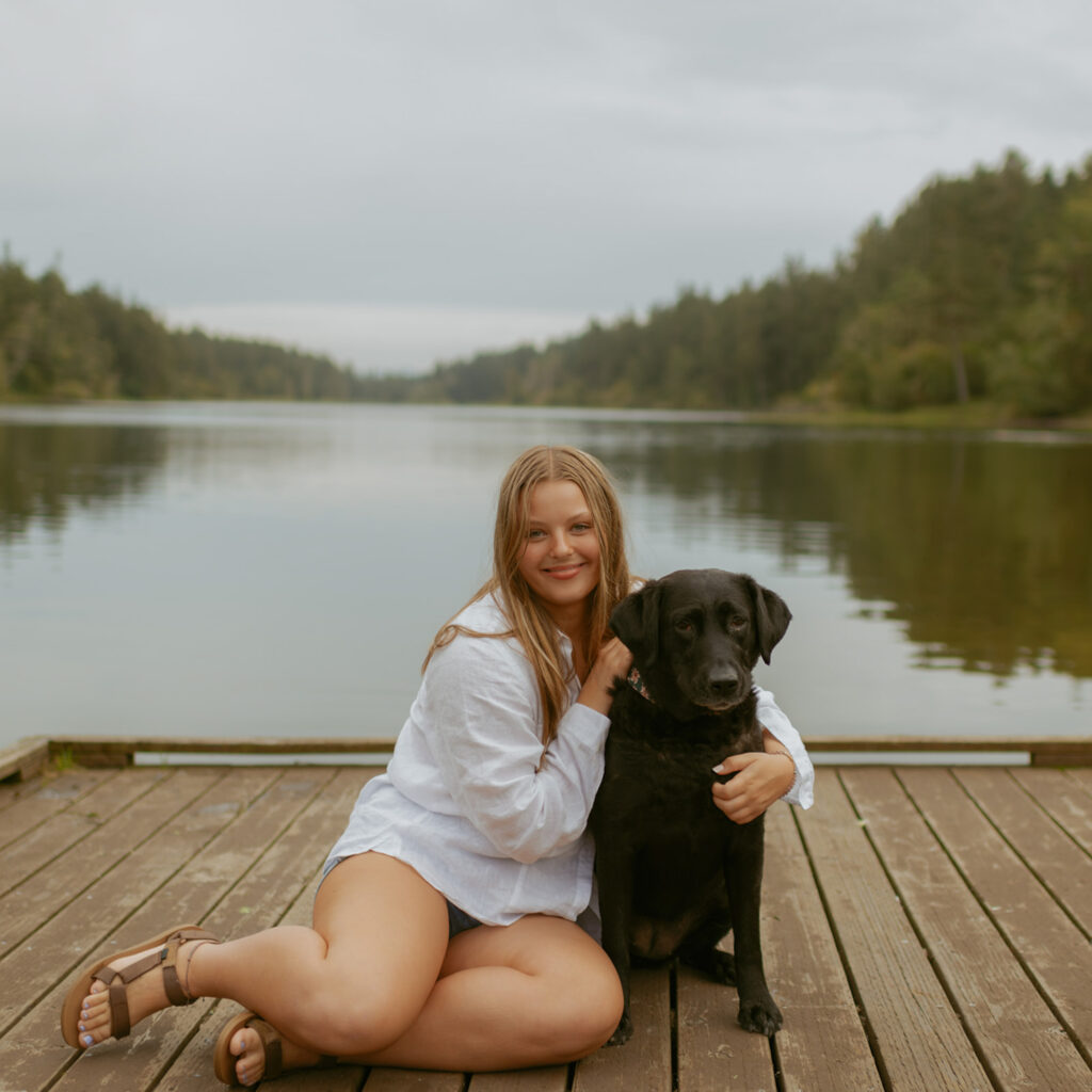 girl sitting in white button up shirt on a dock with her black lab