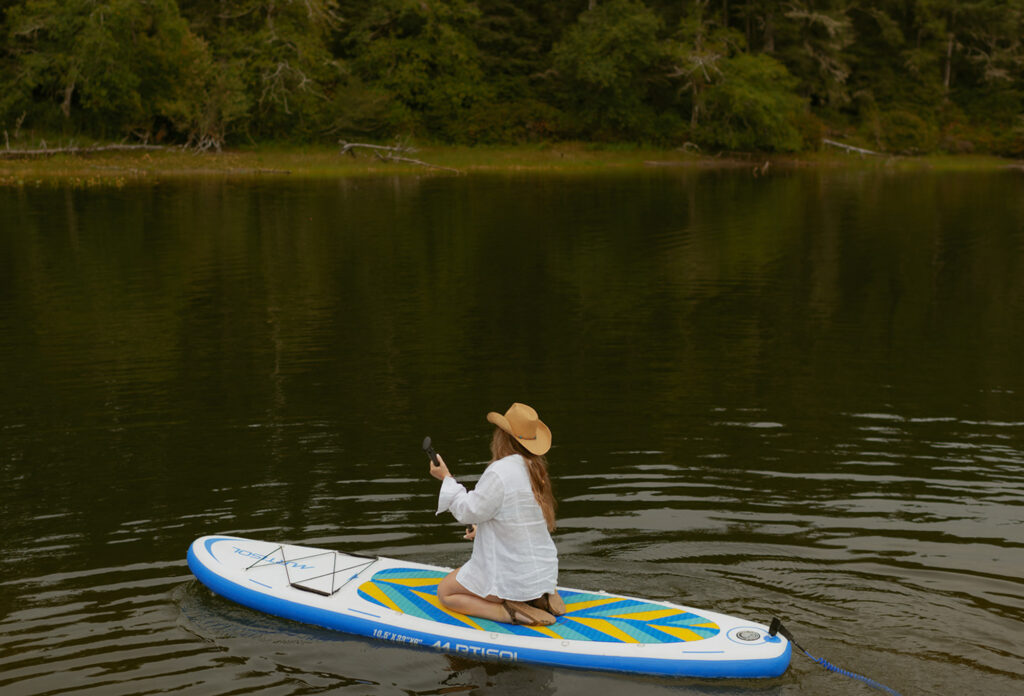 girl paddling across lake on blue and white paddle board. Girl is wearing a cowgirl hat