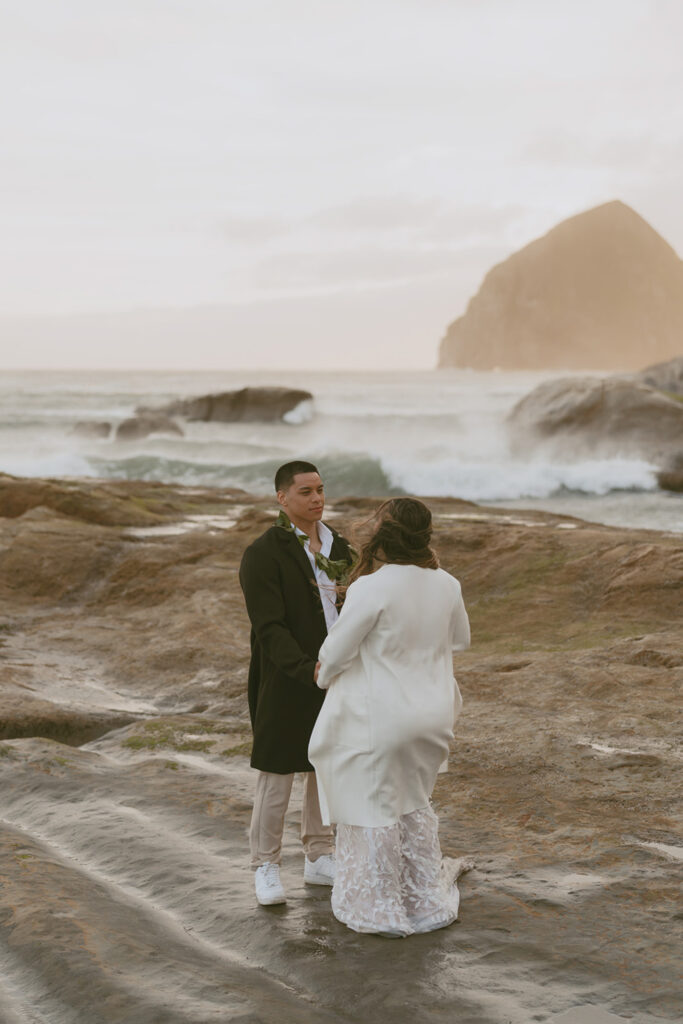 bride and groom facing each other on rocky terrain by the ocean wearing their wedding attire